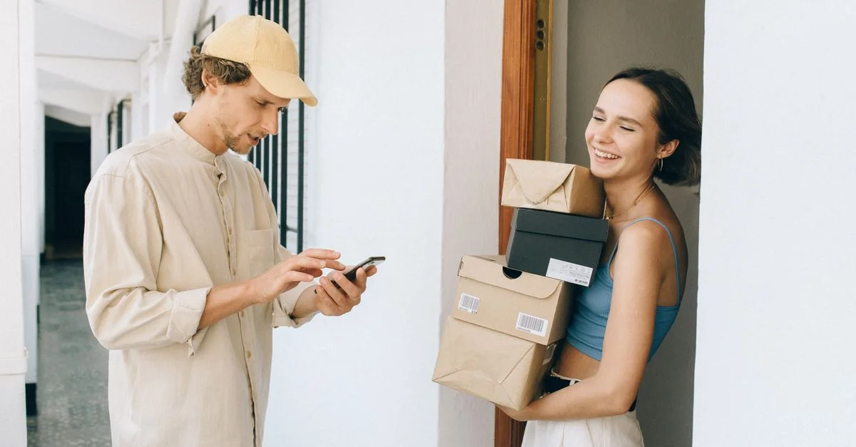 Eine fröhliche Frau erhält eine Lieferung von einer Person in einem Flur und verwendet ein Smartphone für die Online-Zahlung.