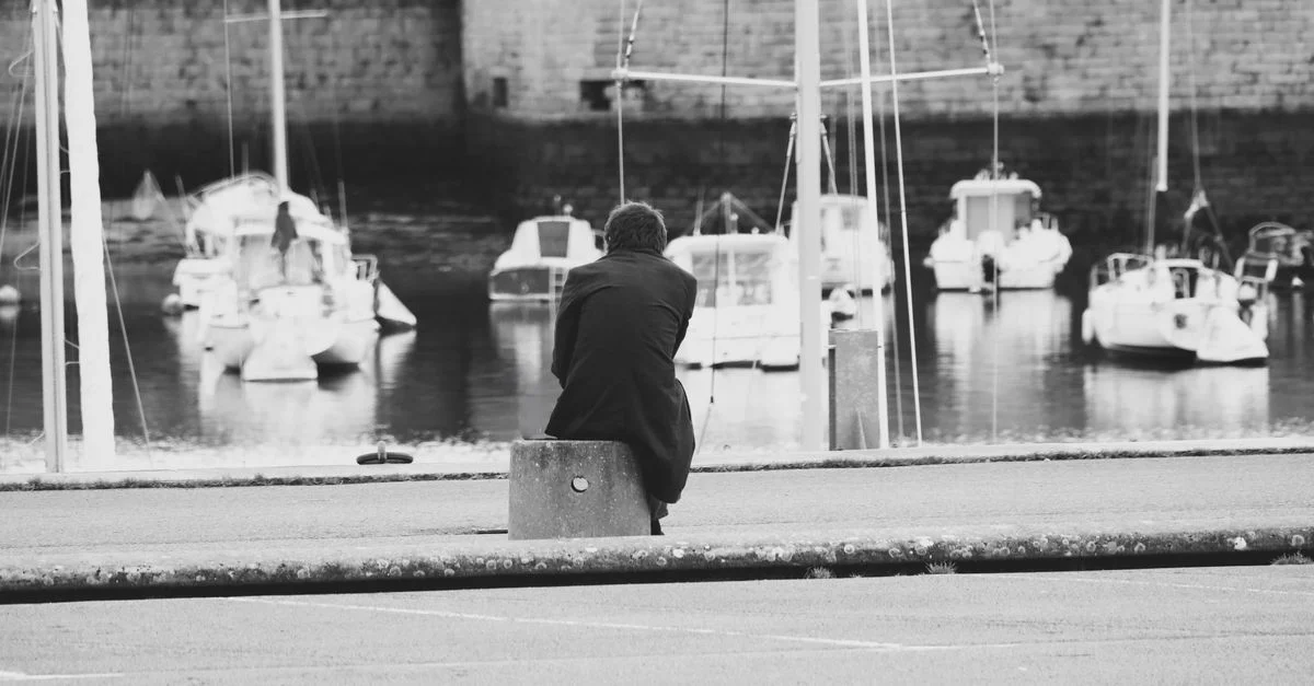 Eine einzelne Person sitzt nachdenklich an einem Hafen und beobachtet in ruhiger Atmosphäre die Boote.