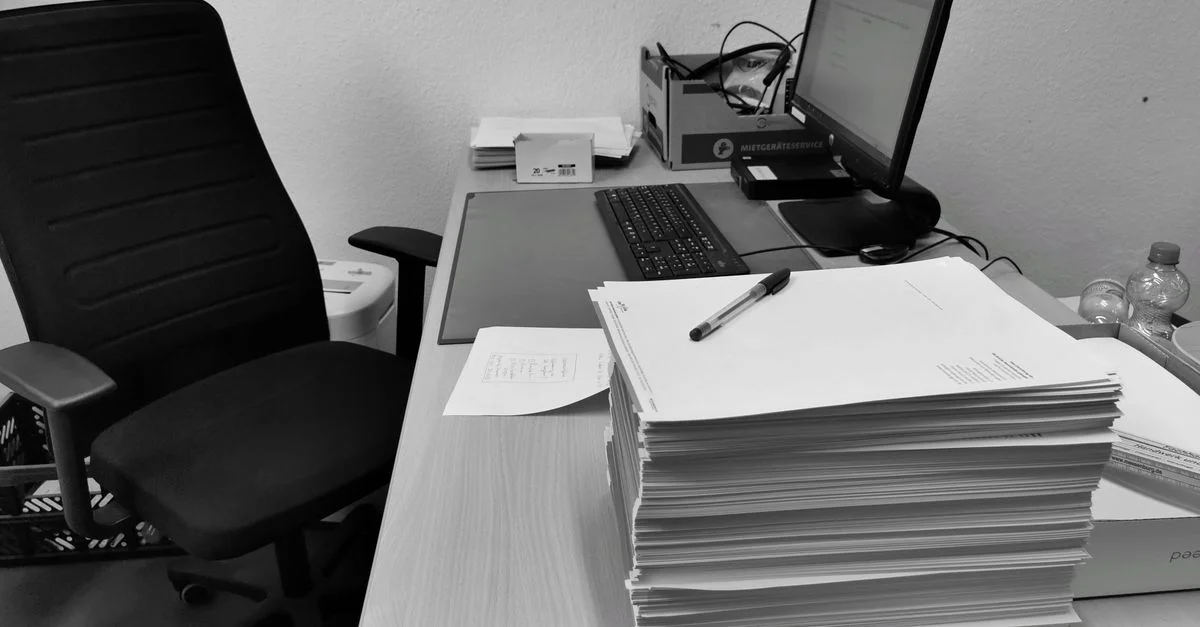 Schwarz-weiße Abbildung eines unordentlichen Schreibtisches mit Computer, Stuhl und Papieren.